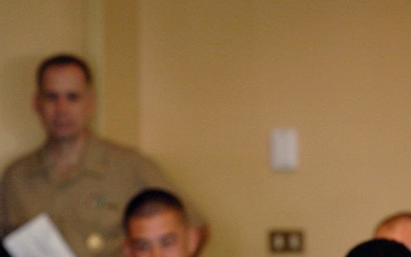 A U.S. Navy servicemember named Joe Campa, wearing a tan/khaki uniform with a name tag and various insignia, is seated at a table and smiling. Several other military personnel in various uniforms are visible in the background, some out of focus. The setting appears to be an indoor dining or meeting event.