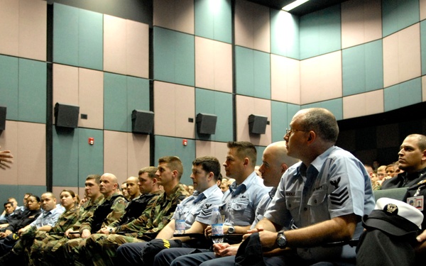 A senior military officer in tan/khaki Navy dress uniform is addressing a seated audience of multi-branch military personnel in an auditorium or briefing room. The audience includes members wearing various uniforms representing multiple U.S. military branches including Navy, Air Force, and Army. The setting appears to be a formal military briefing or training session.