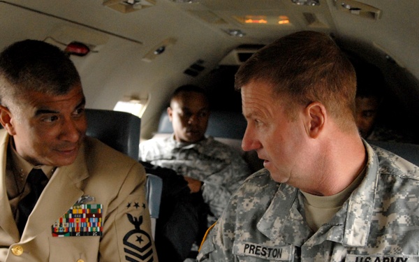 Two senior military personnel are seated inside a small aircraft engaged in conversation. The individual on the right wears U.S. Army ACU (Army Combat Uniform) with the name tape 'PRESTON' and Sergeant Major rank insignia, while the individual on the left wears a U.S. Marine Corps dress uniform with multiple service ribbons and senior enlisted rank insignia. Additional military personnel are visible seated in the background.