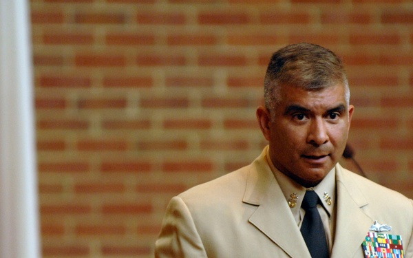 A military officer in a tan/khaki dress uniform is speaking or presenting, likely at a formal military ceremony or briefing. He is gesturing with both hands in a communicative manner, standing in front of a brick wall backdrop. Multiple service ribbons and medals are visible on his left chest.