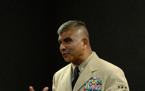 A senior U.S. Navy enlisted leader in Service Dress Khaki uniform is addressing an audience in what appears to be a briefing or meeting room. The individual displays extensive service ribbons, a warfare device, and sleeve insignia consistent with a Master Chief Petty Officer of the Navy (MCPON) or Fleet/Force Master Chief rank. He is gesturing with both hands, suggesting active engagement with the seated audience.