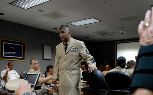 A senior U.S. Navy enlisted officer in Service Dress Khaki uniform stands at the head of a conference table, appearing to lead a briefing or training session. Multiple uniformed Navy personnel are seated around the table, with several raising their hands. The setting is a formal military conference room with overhead lighting, a projection screen, and framed artwork on the wall.