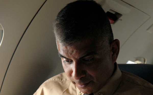 A man in a tan/khaki military uniform with a black tie is seated aboard what appears to be an aircraft, reading documents. He wears collar insignia and a tie tack, both appearing to be gold-colored military devices. The lighting is dramatic, with strong contrast between light and shadow across his face.