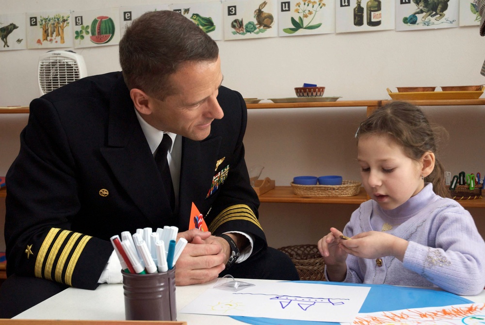 A naval officer in a dark dress uniform with gold sleeve stripes and medals is seated at a table with a young girl in a classroom or educational setting. The officer is attentively watching the child, who appears to be engaged with a small object or craft activity. The setting appears to be a Russian-language classroom based on visible Cyrillic alphabet cards on the wall.
