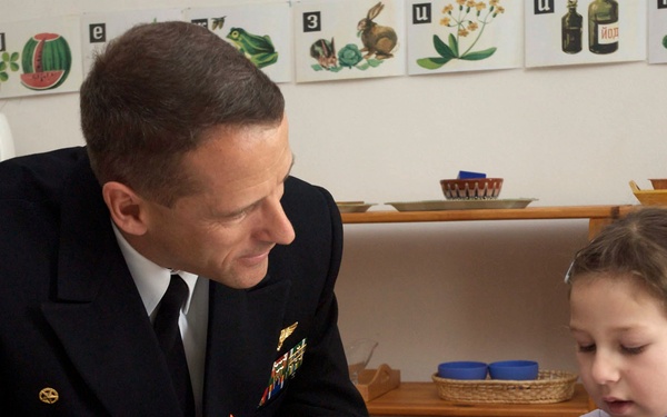 A naval officer in a dark dress uniform with gold sleeve stripes and medals is seated at a table with a young girl in a classroom or educational setting. The officer is attentively watching the child, who appears to be engaged with a small object or craft activity. The setting appears to be a Russian-language classroom based on visible Cyrillic alphabet cards on the wall.