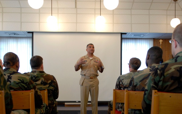 A senior military officer in tan/khaki dress uniform is addressing a group of soldiers seated in rows of wooden chairs in what appears to be a chapel or briefing room. The audience members are wearing woodland camouflage uniforms and are attentively facing the speaker. A projection screen is visible behind the speaker, and a Christian cross is mounted on the wood-paneled wall to the right.