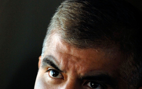 A close-up portrait of a middle-aged male military officer wearing a tan/khaki uniform with multiple service ribbons and insignia visible on the right chest. The dramatic lighting creates strong contrast against a dark background, emphasizing the subject's serious expression. The uniform and decorations suggest a senior non-commissioned or commissioned officer rank.