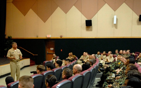A military briefing or address is taking place in an auditorium-style venue. A senior officer in khaki dress uniform stands at the front speaking to a seated audience of military personnel wearing woodland camouflage BDUs. The setting appears to be a formal military theater or lecture hall on a base.