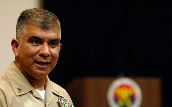 A U.S. Navy officer identified by his name tag as 'Joe Campa' is speaking or presenting at what appears to be a formal military briefing or event. He is wearing a tan/khaki Navy service uniform with multiple ribbons and insignia. A podium with an emblem is visible in the blurred background.