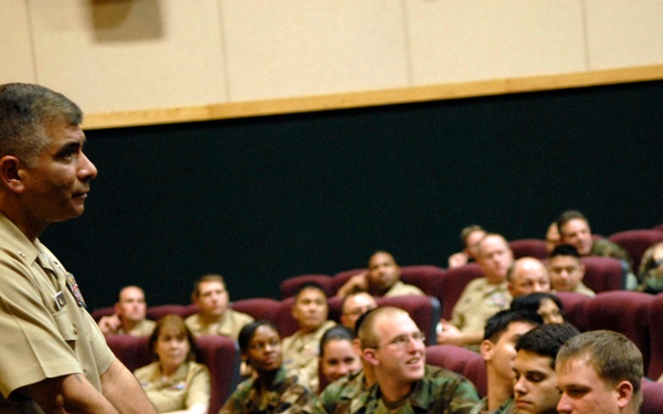 A senior military officer in tan/khaki service uniform is addressing a seated audience of military personnel in an auditorium or briefing room. The audience members are wearing a mix of camouflage battle dress uniforms and tan service uniforms, suggesting a mixed-branch or multi-unit gathering. The officer appears to be delivering a briefing, lecture, or command address.