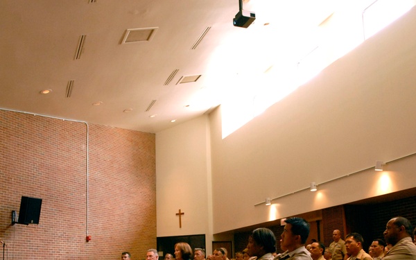A military officer in a tan/khaki dress uniform is addressing a seated audience of fellow military personnel in what appears to be a chapel or multi-purpose religious facility, evidenced by the visible Christian cross on the wall. The speaker stands at a wooden podium on a raised platform, gesturing with a thumbs-up or pointing gesture toward the audience. The audience consists of approximately 30-40 uniformed military personnel seated in wooden chairs with green cushions.