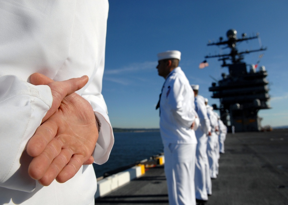 A line of U.S. Navy sailors in white dress uniforms are standing at parade rest on the flight deck of an aircraft carrier. The sailors are positioned in a formal formation, facing outward toward the water. A large ship's island superstructure with radar equipment and a visible American flag is seen in the background.