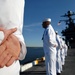 A line of U.S. Navy sailors in white dress uniforms are standing at parade rest on the flight deck of an aircraft carrier. The sailors are positioned in a formal formation, facing outward toward the water. A large ship's island superstructure with radar equipment and a visible American flag is seen in the background.