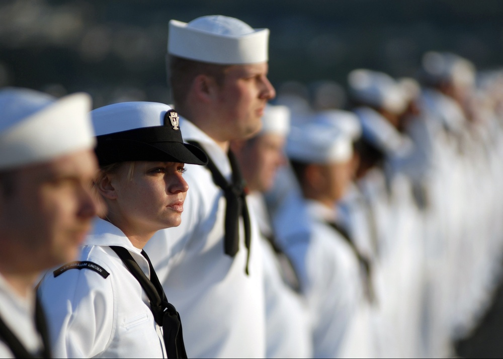 A formation of U.S. Navy sailors dressed in white dress uniforms are standing at attention in a disciplined line. The focal point is a female sailor in the foreground wearing a combination cover (white cap with black brim) and a black neckerchief. A name tape reading 'USS Abraham Lincoln' is visible on her uniform, suggesting this is a naval ceremony or formal muster.