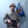 A young male airman in full combat gear stands at attention in what appears to be an indoor military facility. He is wearing US Air Force Airman Battle Uniform (ABU) with body armor, helmet, and holding an M4 carbine rifle. A unit guidon flag is visible behind him, partially readable as '586 ELRS' with Air Force insignia.