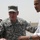 A U.S. Army general in ACU (Army Combat Uniform) with a name tape reading 'PETRAEUS' and four-star rank insignia is engaged in conversation with a civilian man in a white shirt. The setting appears to be an outdoor military installation in an arid environment. Additional military personnel are visible in the background.