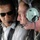 Two individuals are seated inside what appears to be a military helicopter or aircraft. Both are wearing aviation-style headsets with microphones. One person is wearing a white dress shirt with a safety harness and sunglasses, while the other is wearing military camouflage (ACU pattern) uniform.