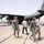 The image shows military personnel and civilians on an airfield tarmac near a large gray military transport aircraft. The aircraft displays tail markings including 'AMC', 'DYESS', and the number '41663', along with a US flag and Texas state flag insignia. Several individuals in US Army combat uniforms are walking alongside civilians on the flight line.