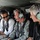 Three individuals are seated inside a military helicopter, all wearing aviation headsets. One person is dressed in civilian attire (white shirt and tie with sunglasses), one is in military camouflage uniform with a Ranger tab visible, and the third wears a black polo shirt. The group appears to be engaged in conversation, with at least one person smiling.