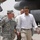 The image shows a military officer in U.S. Army ACU (Army Combat Uniform) walking alongside a man in civilian attire (white shirt and khaki pants) on what appears to be a military airfield or tarmac. A large military aircraft is visible in the background along with military vehicles. The name tape on the uniformed officer reads 'PETRAEUS' and four-star rank insignia is visible.