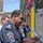 An Iraqi Police officer with three-star rank insignia is cutting a ribbon with scissors, indicating a formal opening or dedication ceremony. Several other uniformed personnel are visible in the background, one of whom appears to be photographing the event. Colorful decorative flowers and streamers adorn the entrance structure, consistent with a celebratory occasion.