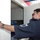 A uniformed security or law enforcement officer is inspecting the interior of a large white security scanning machine, likely an X-ray baggage scanner. The officer is wearing a dark uniform with a shoulder patch and a wristwatch, and is peering into the machine's opening. The setting appears to be a security checkpoint, possibly at an airport, border crossing, or public venue.