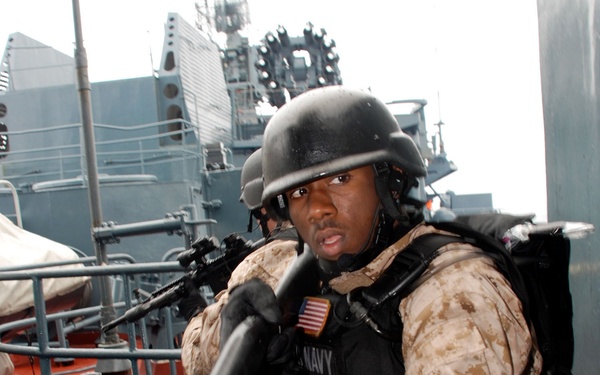 A U.S. Navy sailor in full tactical gear is positioned on the deck of a naval vessel, holding what appears to be a shotgun or breaching tool. The sailor is wearing desert camouflage (MARPAT desert pattern), a ballistic helmet, body armor with a 'NAVY' label, and an American flag patch on the left shoulder. A second armed sailor is visible in the background, and a large gray warship is visible behind them.