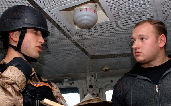 A U.S. military personnel in full combat gear, including a helmet, body armor, and desert camouflage uniform, appears to be conducting an inspection or interview aboard a vessel. They are holding an open document or logbook while speaking with a civilian man wearing a gray 'Lamzar' branded hoodie. The setting appears to be the interior of a ship's bridge or cabin.