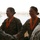 Two female U.S. Army soldiers wearing digital camouflage uniforms are being congratulated and are receiving medals with orange ribbons. A senior soldier on the left is shaking hands with one of the recipients. The setting appears to be an outdoor or semi-open structure with a body of water visible in the background.