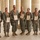 Seven U.S. Army soldiers in ACU (Army Combat Uniform) camouflage are posed together in a formal group photo at what appears to be a colonnaded building. Each soldier is wearing an orange medal ribbon around their neck and holding a framed certificate or award plaque. This appears to be an awards ceremony or recognition event.