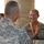 A U.S. Army female soldier named Smith, wearing an orange medal/lanyard, is smiling while receiving what appears to be an award or recognition plaque from a senior male soldier. The ceremony appears to be taking place on an outdoor terrace or balcony with a body of water visible in the background. Both individuals are wearing Army Combat Uniforms (ACU) in the digital camouflage pattern.