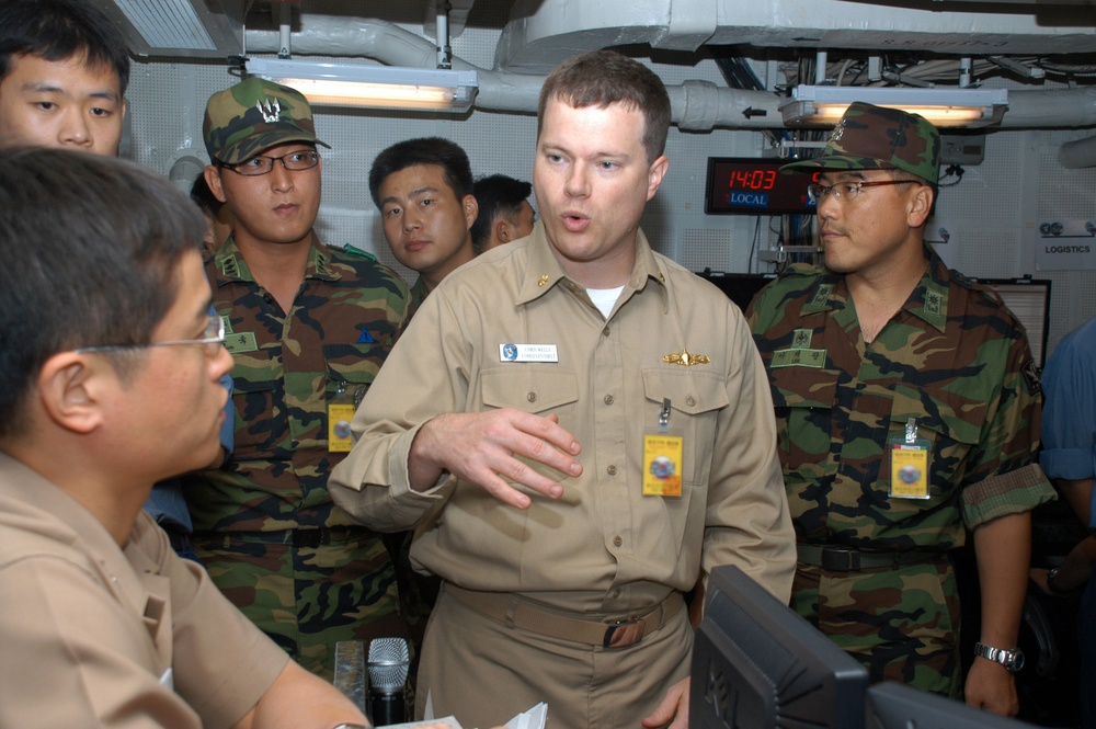 A U.S. Navy officer in tan/khaki uniform is briefing or explaining something to a group of military personnel wearing woodland camouflage uniforms, likely South Korean military members, in what appears to be a shipboard operations or logistics room. The American officer is gesturing with his hand while speaking, and the visitors are wearing yellow visitor badges. A digital clock on the wall displays 14:03 LOCAL time.