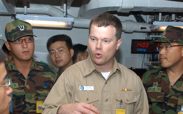 A U.S. Navy officer in tan/khaki uniform is briefing or explaining something to a group of military personnel wearing woodland camouflage uniforms, likely South Korean military members, in what appears to be a shipboard operations or logistics room. The American officer is gesturing with his hand while speaking, and the visitors are wearing yellow visitor badges. A digital clock on the wall displays 14:03 LOCAL time.