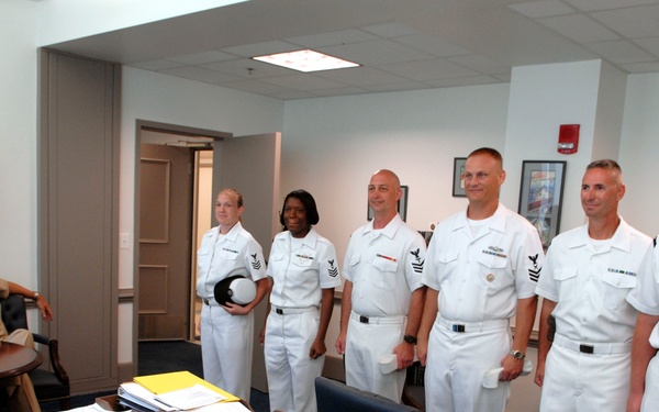 A senior naval officer in khaki uniform addresses a group of six enlisted personnel in white dress uniforms standing in a line inside what appears to be a military office. The setting includes a large desk covered with documents, binders, and papers, along with framed artwork on the walls suggesting a command office environment. The scene appears to be a formal meeting, briefing, or recognition ceremony.