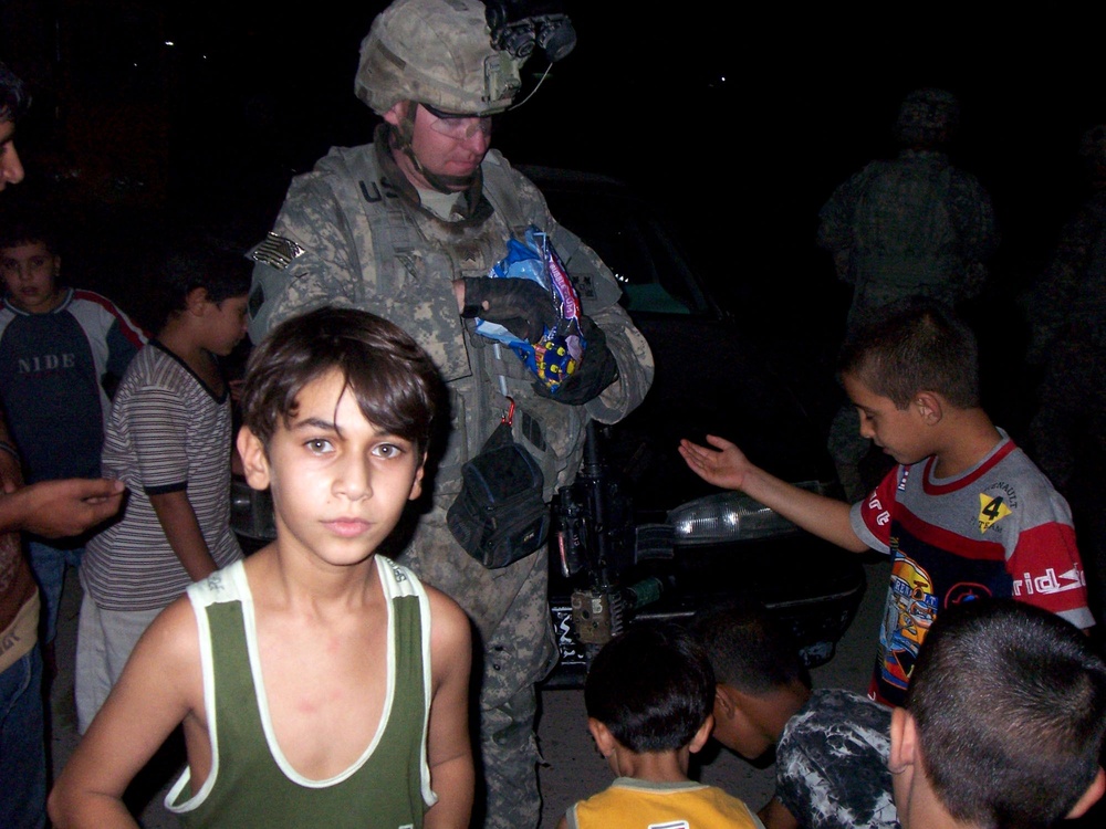 A U.S. Army soldier in full combat gear, including a helmet with night-vision mount and ACU camouflage uniform, is surrounded by a group of children at night. The soldier appears to be distributing snacks or candy from a colorful bag to the children gathered around. A second soldier is visible in the dark background near a vehicle.