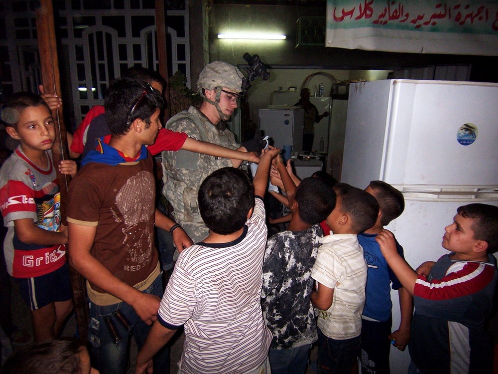 A U.S. military soldier in combat uniform and helmet is surrounded by a group of children and young people in what appears to be an indoor setting at night. The soldier appears to be distributing or showing something small to the gathered crowd. Arabic text is visible on a banner in the background, suggesting a Middle Eastern location.