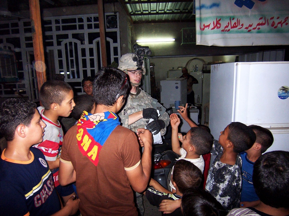 A U.S. Army soldier in full combat gear, including helmet with night-vision device mount, is surrounded by a group of local children in what appears to be an indoor or semi-covered civilian space at night. The soldier appears to be interacting positively with the children, who are crowding around with apparent curiosity and enthusiasm. Arabic text is visible on a banner in the background, and the setting appears to be in a Middle Eastern country.