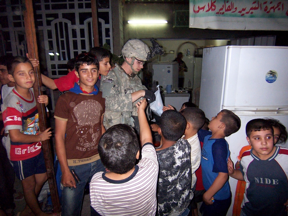 A uniformed military soldier wearing combat fatigues and a helmet is surrounded by a large group of children in what appears to be an indoor setting in the Middle East. The soldier appears to be distributing items from a plastic bag to the eager children gathered around. A banner with Arabic text is visible in the background, along with a refrigerator and interior furnishings.