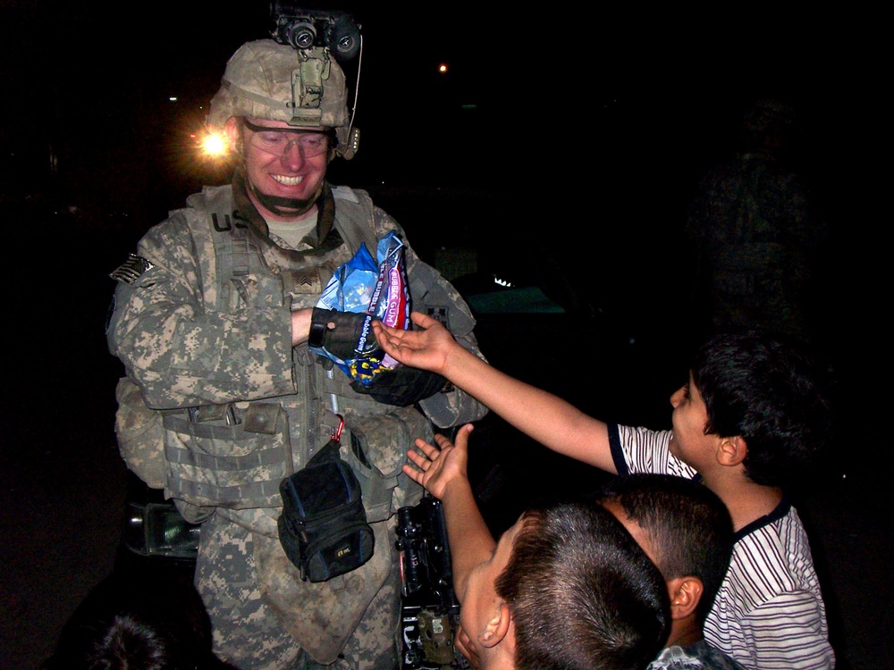 A U.S. Army soldier in full combat gear, including ACU (Army Combat Uniform) and helmet with night vision mount, is distributing candy/bubble gum to local children at night. The soldier is smiling warmly while children reach out to receive the treats. The scene appears to depict a civil affairs or community engagement interaction in a deployed environment.