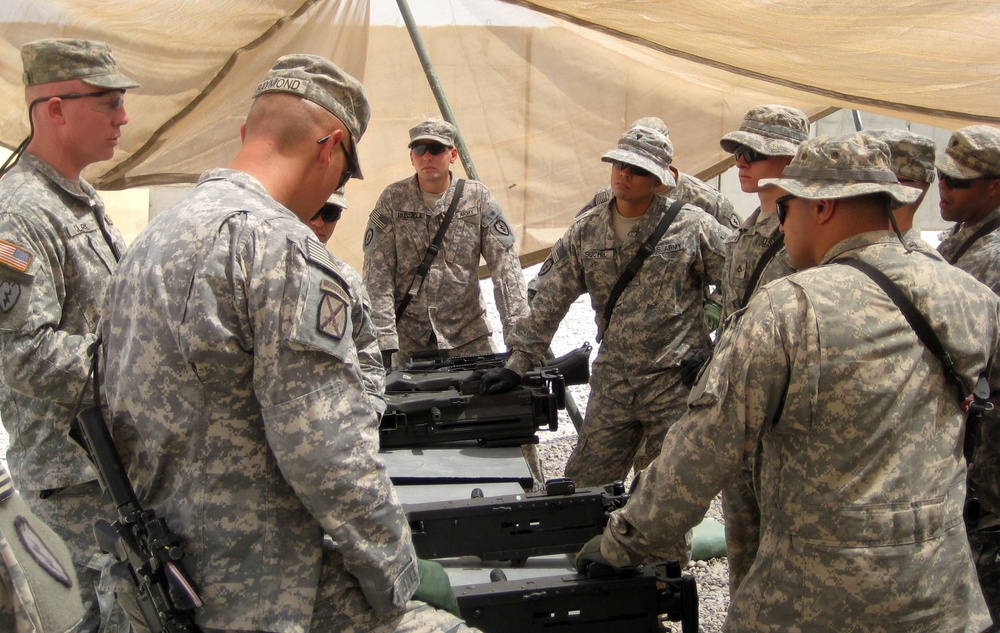 A group of U.S. Army soldiers in ACU (Army Combat Uniform) digital camouflage are gathered under a tan shade canopy, appearing to conduct a training or equipment familiarization session. They are clustered around what appears to be a weapons system or military equipment laid out on a surface. The setting appears to be a forward operating base or training area in an arid environment.