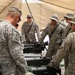 A group of U.S. Army soldiers in ACU (Army Combat Uniform) digital camouflage are gathered under a tan shade canopy, appearing to conduct a training or equipment familiarization session. They are clustered around what appears to be a weapons system or military equipment laid out on a surface. The setting appears to be a forward operating base or training area in an arid environment.