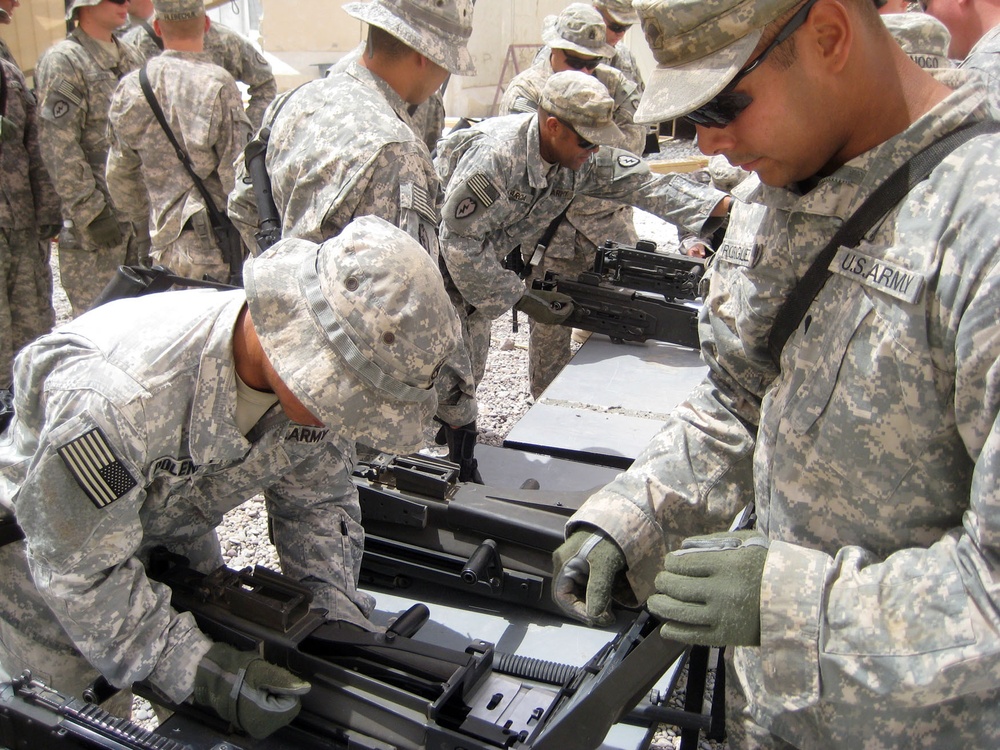 A group of U.S. Army soldiers in ACU (Army Combat Uniform) camouflage are engaged in weapons maintenance or assembly training outdoors. Multiple soldiers are working on what appears to be a heavy machine gun, likely an M2 .50 caliber or M240, laid out on a flat surface. The setting appears to be a forward operating base or training area in an arid environment.