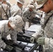 A group of U.S. Army soldiers in ACU (Army Combat Uniform) camouflage are engaged in weapons maintenance or assembly training outdoors. Multiple soldiers are working on what appears to be a heavy machine gun, likely an M2 .50 caliber or M240, laid out on a flat surface. The setting appears to be a forward operating base or training area in an arid environment.