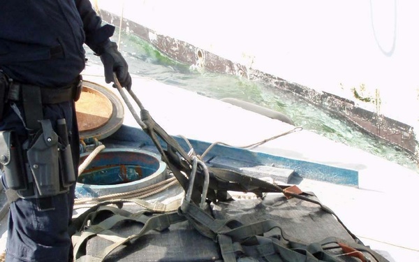 The image shows what appears to be a law enforcement or coast guard interdiction operation aboard a vessel. A uniformed officer armed with a holstered sidearm is handling a large bundle of tightly wrapped packages secured with black strapping and orange ratchet straps, consistent with contraband seizure operations. The setting appears to be the deck of a ship or boat near open water.