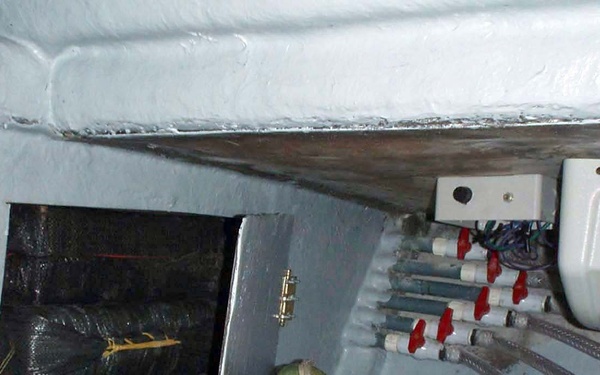 The image shows a cramped interior space, likely the bilge or engine compartment area of a boat or vessel. The space contains plumbing manifolds with red ball valves, electrical panels, flexible hoses, and various stored items. The area appears utilitarian and cluttered, consistent with a marine vessel's below-deck utility space.
