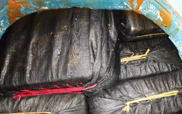 The image shows the interior of a circular hatch or access port, likely on a vessel or industrial container, revealing tightly packed black mesh/woven bags secured with colored ties. The bags appear wet or oily and are densely stacked to fill the circular opening. This type of concealment inside a vessel compartment is consistent with documented smuggling interdiction photography.