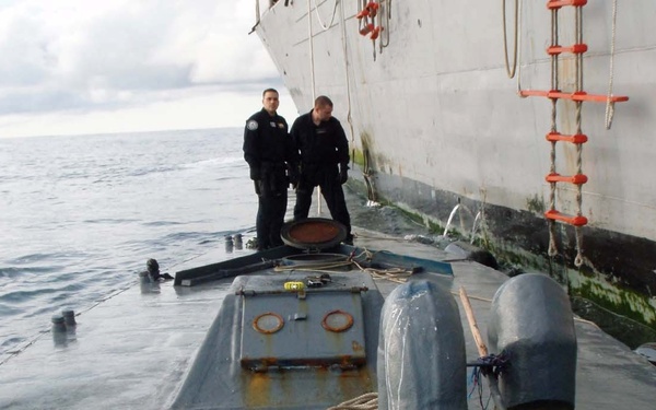 Two law enforcement officers in dark uniforms are standing on the deck of a low-profile semi-submersible vessel (narco submarine) alongside a larger ship. The semi-submersible appears to have been intercepted at sea, with its hatch open and various items visible on its deck. This appears to be a maritime interdiction operation, likely a drug trafficking seizure.