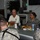 A civilian in a white polo shirt with an emblem is seated at a table having a meal with several U.S. Army soldiers in ACU (Army Combat Uniform) camouflage. The setting appears to be a military dining facility or conference room, with food trays, water bottles, and name placards visible on the table. The civilian appears to be speaking or briefing the soldiers during the meal.