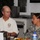 Two individuals are seated at a table in what appears to be a military dining facility or meeting room. One person is wearing a white polo shirt with a Department of Defense 'Enduring Freedom' Pentagon emblem, while the other is a U.S. Army soldier in ACU (Army Combat Uniform) with a name tape partially readable as 'BODONIAN' or similar. They appear to be engaged in conversation over a meal.