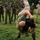 A military combatives or hand-to-hand combat training demonstration is taking place on a grass field. One person in a green t-shirt is executing a throw or takedown on another individual in camouflage uniform, while a group of uniformed soldiers looks on. The observing group includes personnel in two distinct camouflage patterns, suggesting a joint or multinational military training exercise.